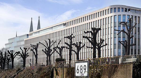 Im Zentrum des Bildes steht ein großes weißes Gebäude, in dem das Institut der Deutschen Wirtschaft in Köln untergebracht ist. Im Vordergrund stehen beschnittene Bäume direkt am Rheinufer. Eine Flusskilometer-Tafel mit der Zahl 689 ist auch zu sehen. Hinter dem Gebäude ragen die beiden Türme des Kölner Domes hervor.