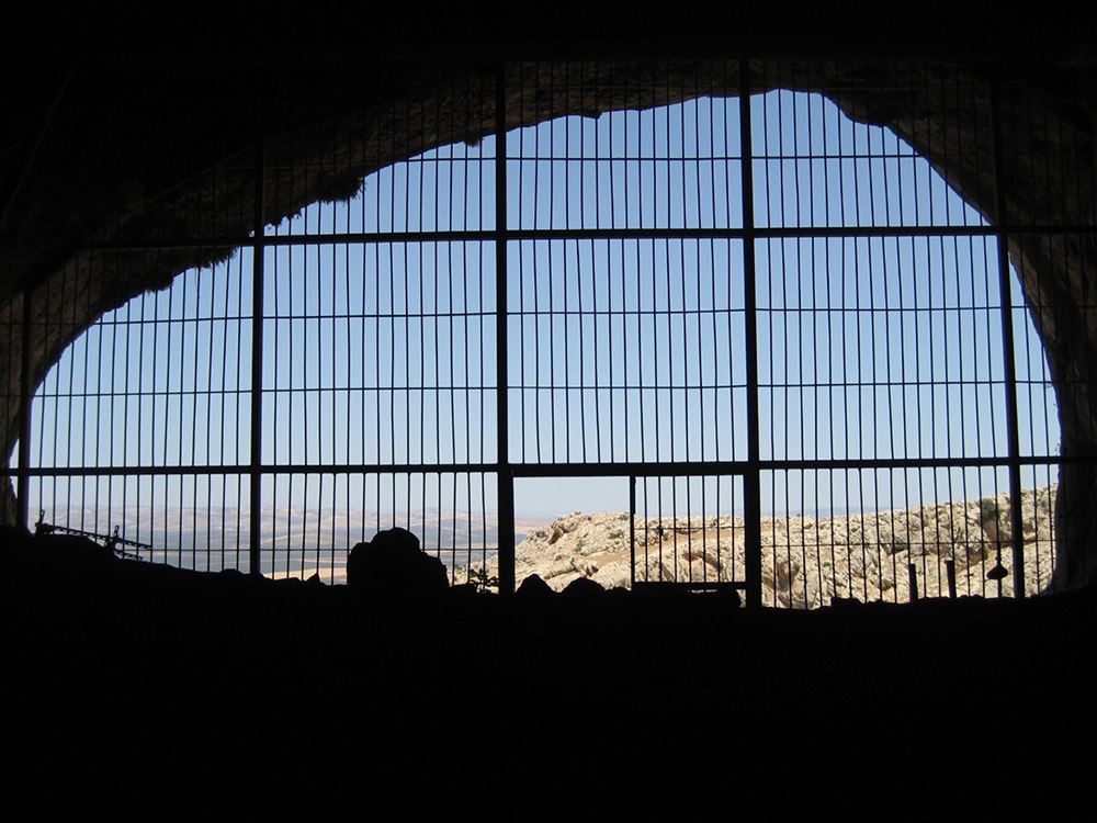 Blick aus einer Höhle durch ein engmaschiges Gitter auf eine Landschaft mit Gebäuden.