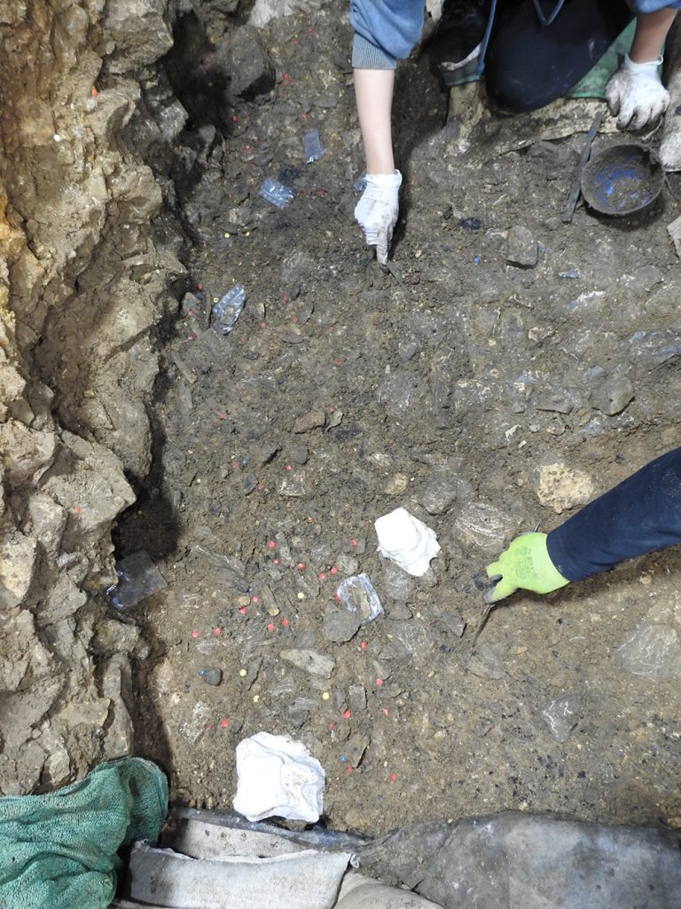 Eine Grabungsstelle mit vielen Steinen und Scherben. Zwei Hände mit Handschuhen und Werkzeugen bei der Grabungsarbeit.