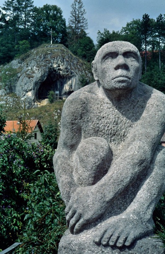 Eine Steinskulptur in Form eines Neandertales steht vor einem Höheneingang, Daneben orangene Blumen.