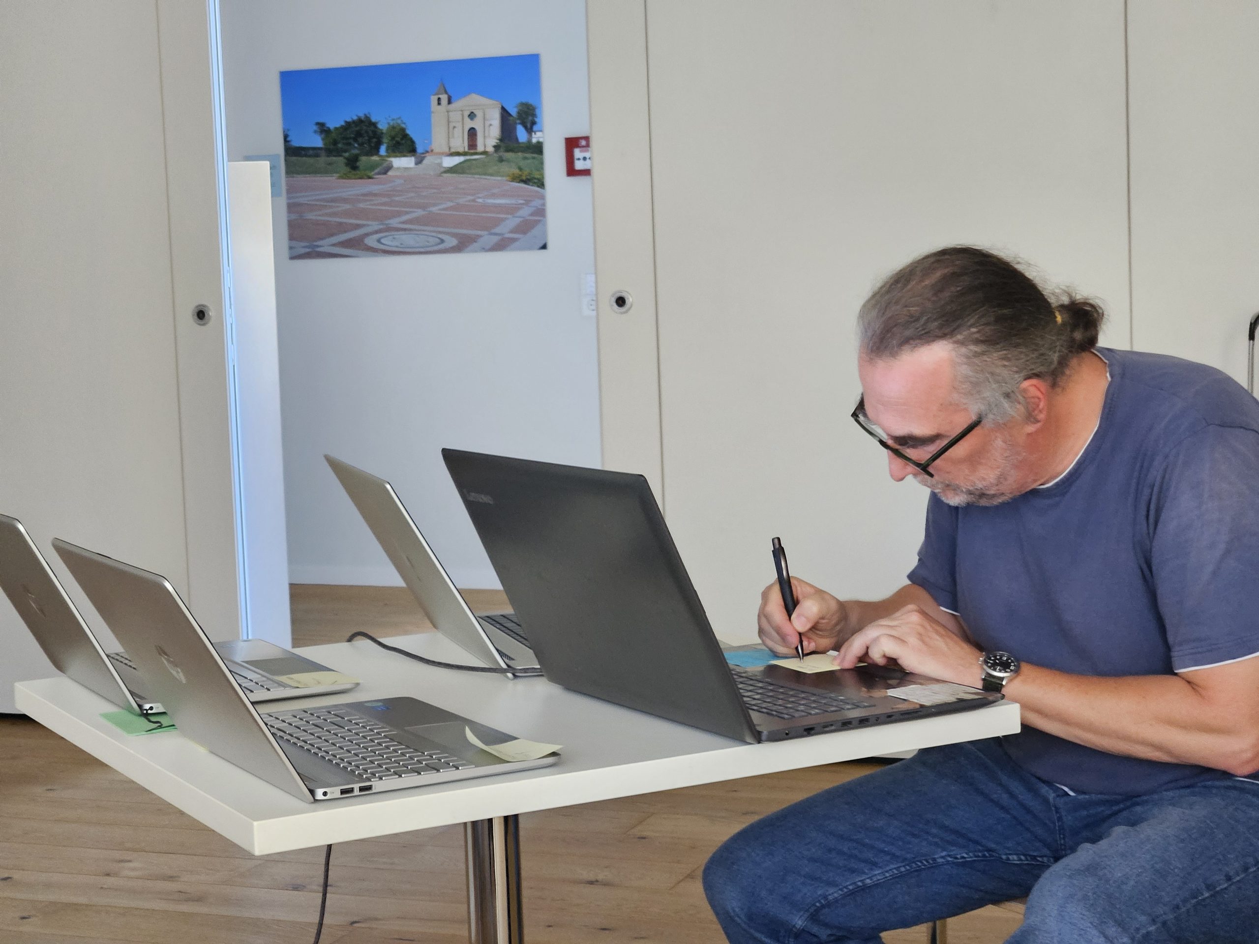 Ein Mann hat einen Kugelschreiber in der Hand. Vor ihm stehen vier aufgeklappte Laptops. Er schreibt aber mit dem Kugelschreiber.