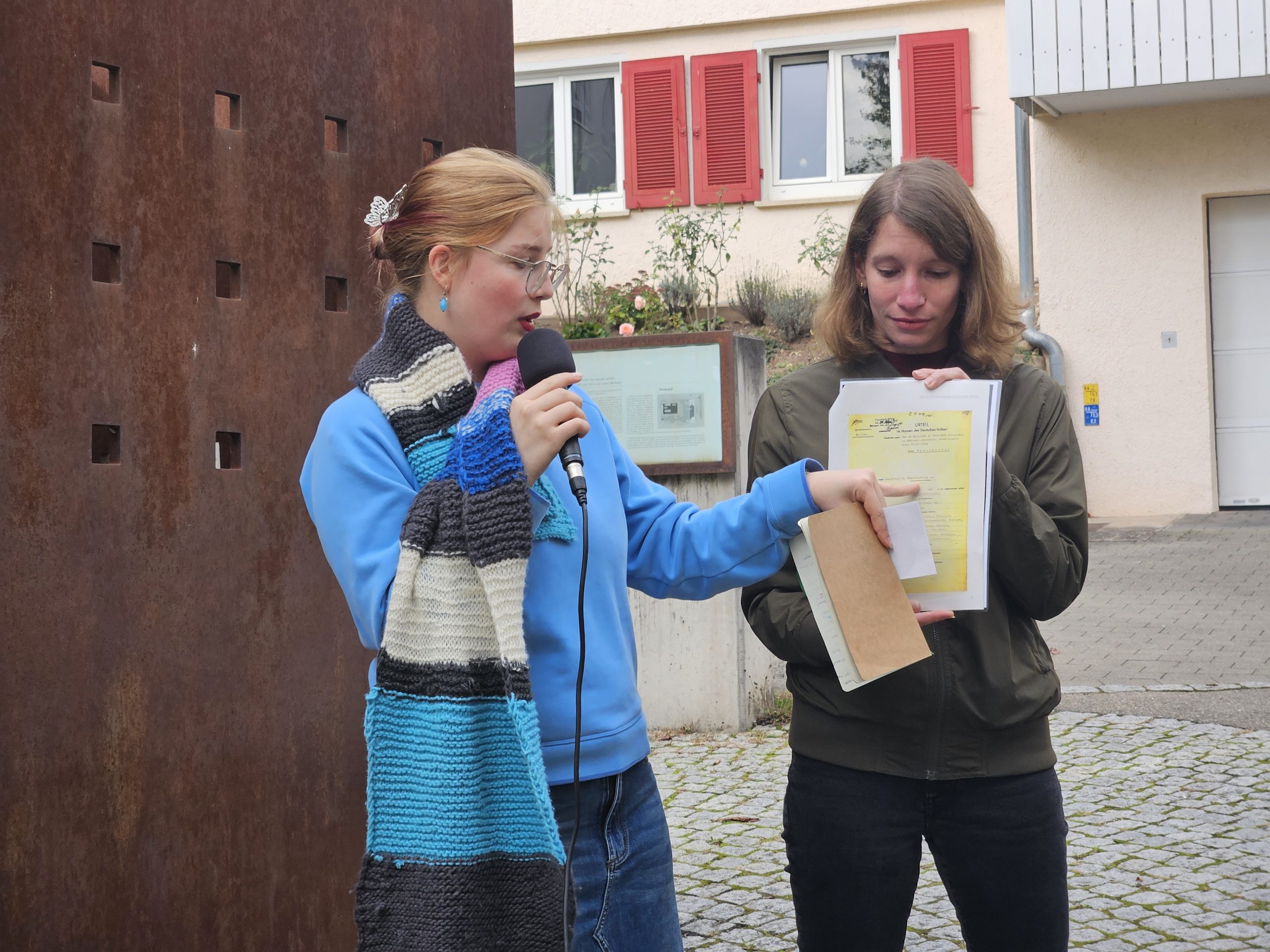 Zwei junge Frauen stehen vor einer verrosteten Eisenwand mit kleinen quadratischen Aussägungen. Es ist das Denkmal am Tübinger Synagogenplatz. Sie halten Fotokopien von historischen Dokumenten in der Hand.