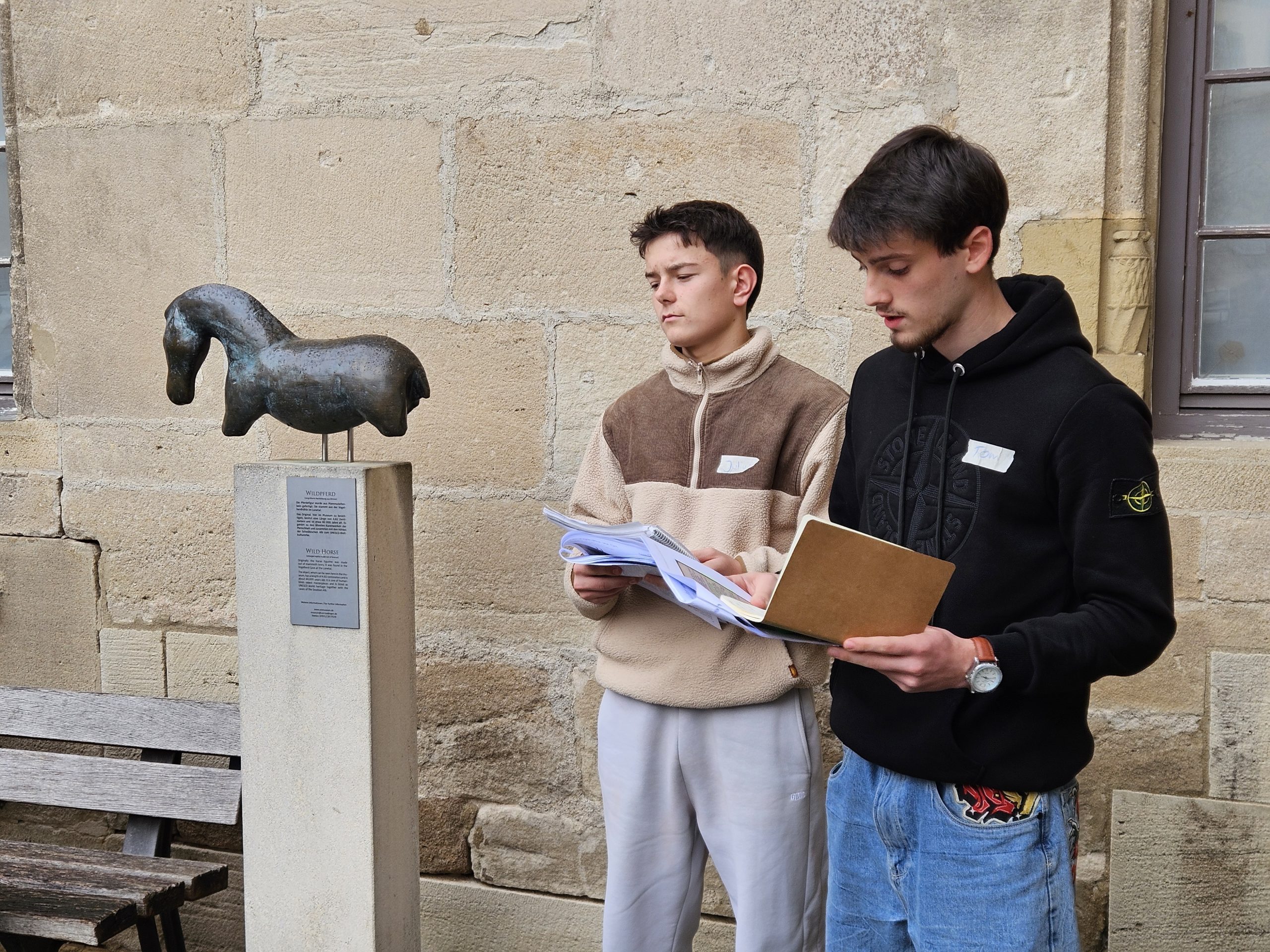Neben einem vergrößerten Bronzeabguss einer Pferdefigur von der Vogelherdhöhle im Innenhof des Schlosses Hohentübingen stehen zwei junge Männer, die ausdedruckte Schriftstücke in der Hand halten.
