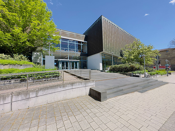 Various buildings of the University of Tübingen, trees and bushes next to them, a paved walkway in the foreground.