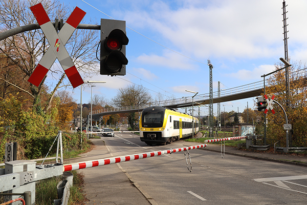 Ein gelb-weiß-schwarzer Triebwagen fährt an einem beschrankten Bahnübergang über die Straße.