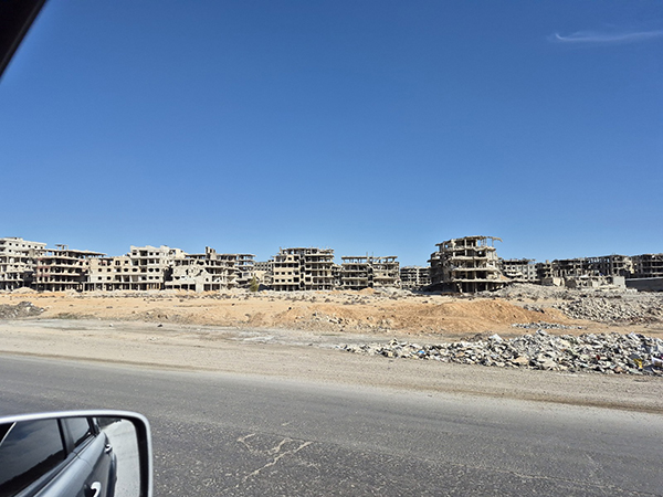 Blick von einer Straße über trockenes, wüstenhaftes Land auf vom Kriegs zerstörte Häuser und Wohnblocks.