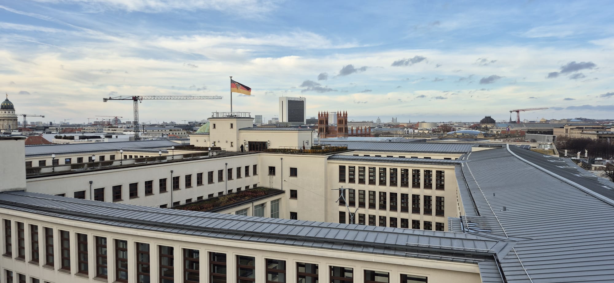 Blick über das Auswärtige Amt in Berlin über die Dächer Berlins mit zwei Baukränen und einer wehenden Deutschland-Flagge.