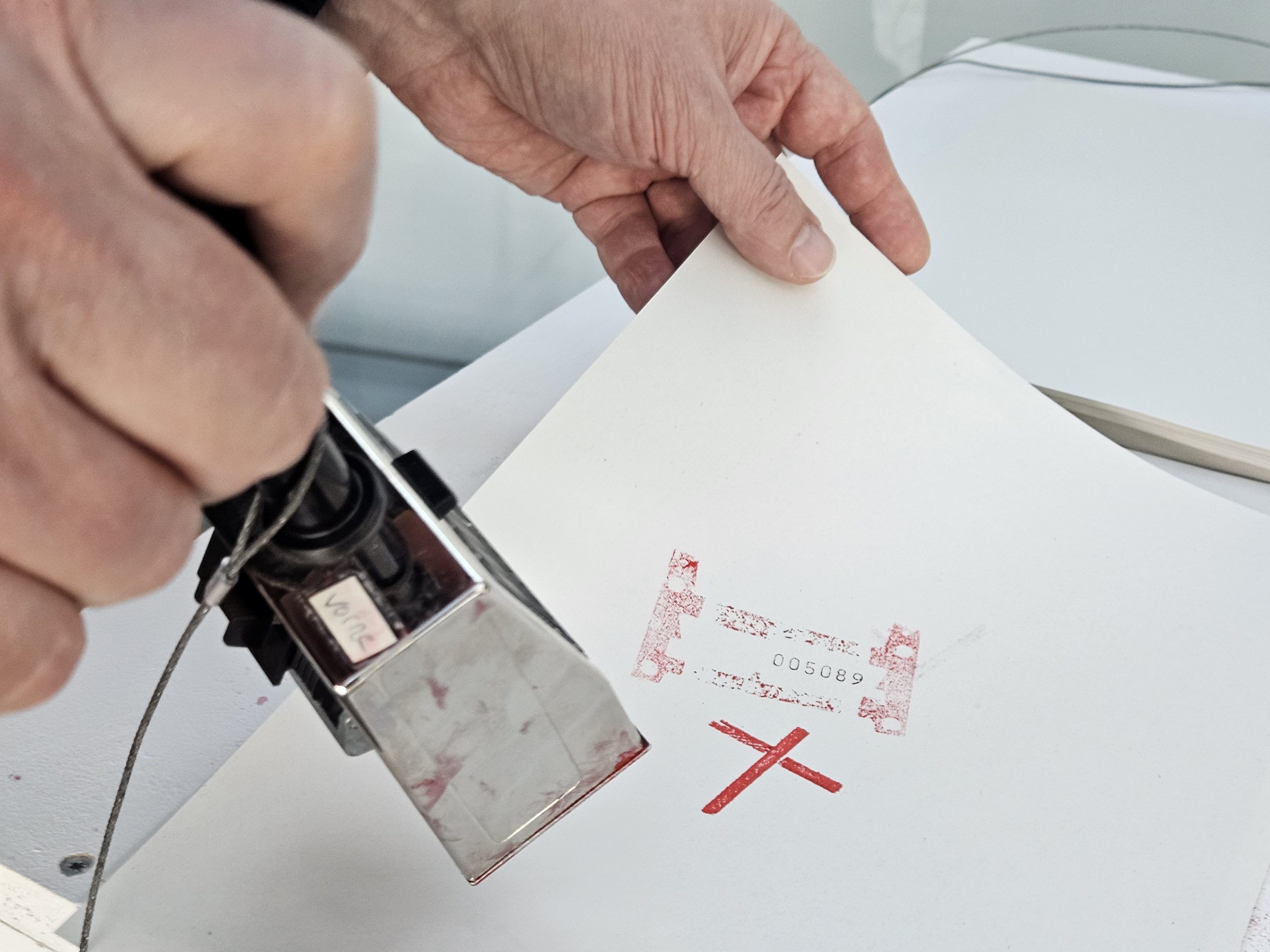 Das Foto zeigt eine Hand, die einen Stempel führt. Auf einem Papierblatt sind zwei Stempel zu sehen, einer mit einem roten Kreuz und einer mit einer Nummer.